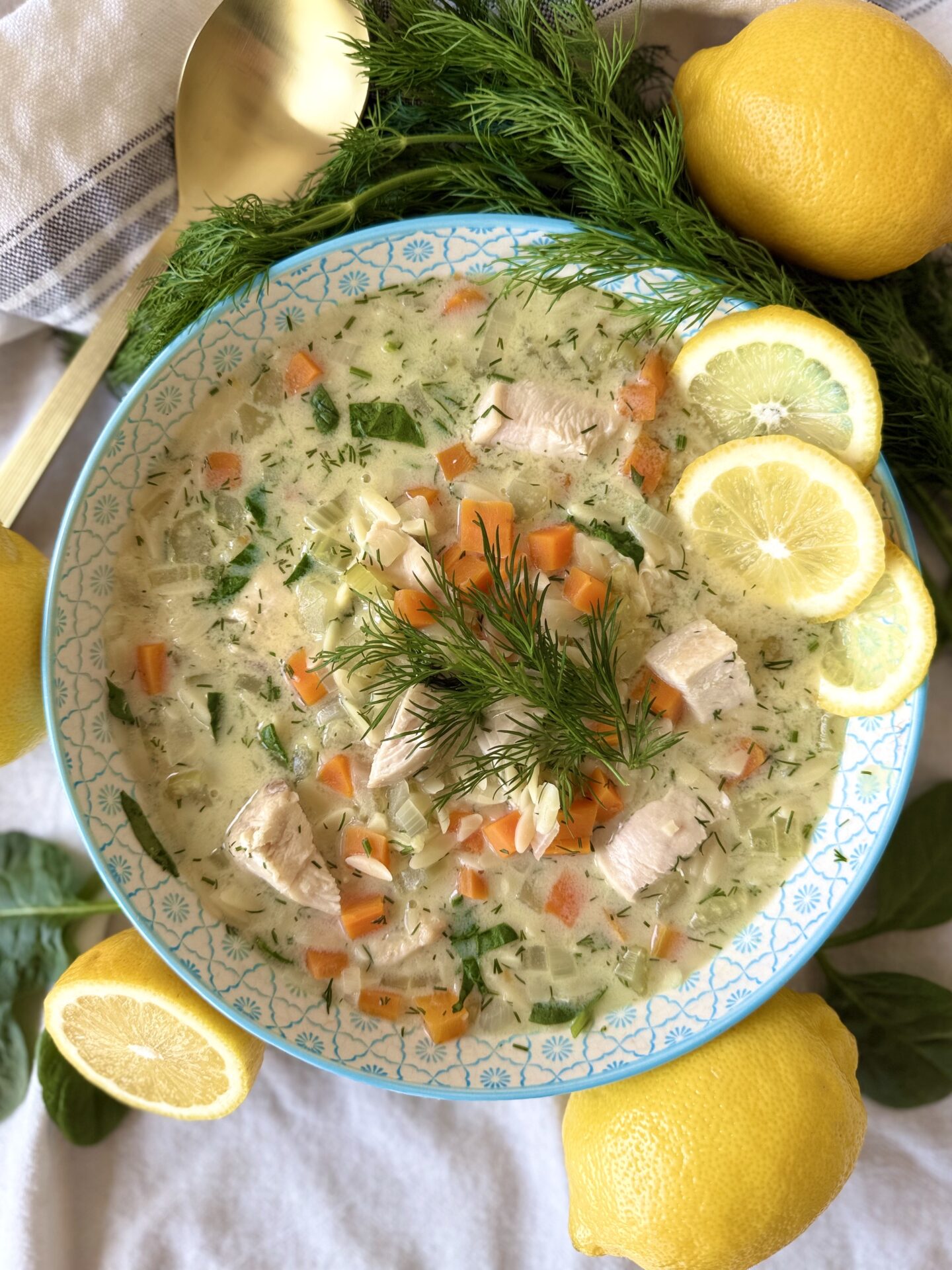 A blue and white bowl filled with Avgolemono soup is seen from above surounded by lemons and dill.