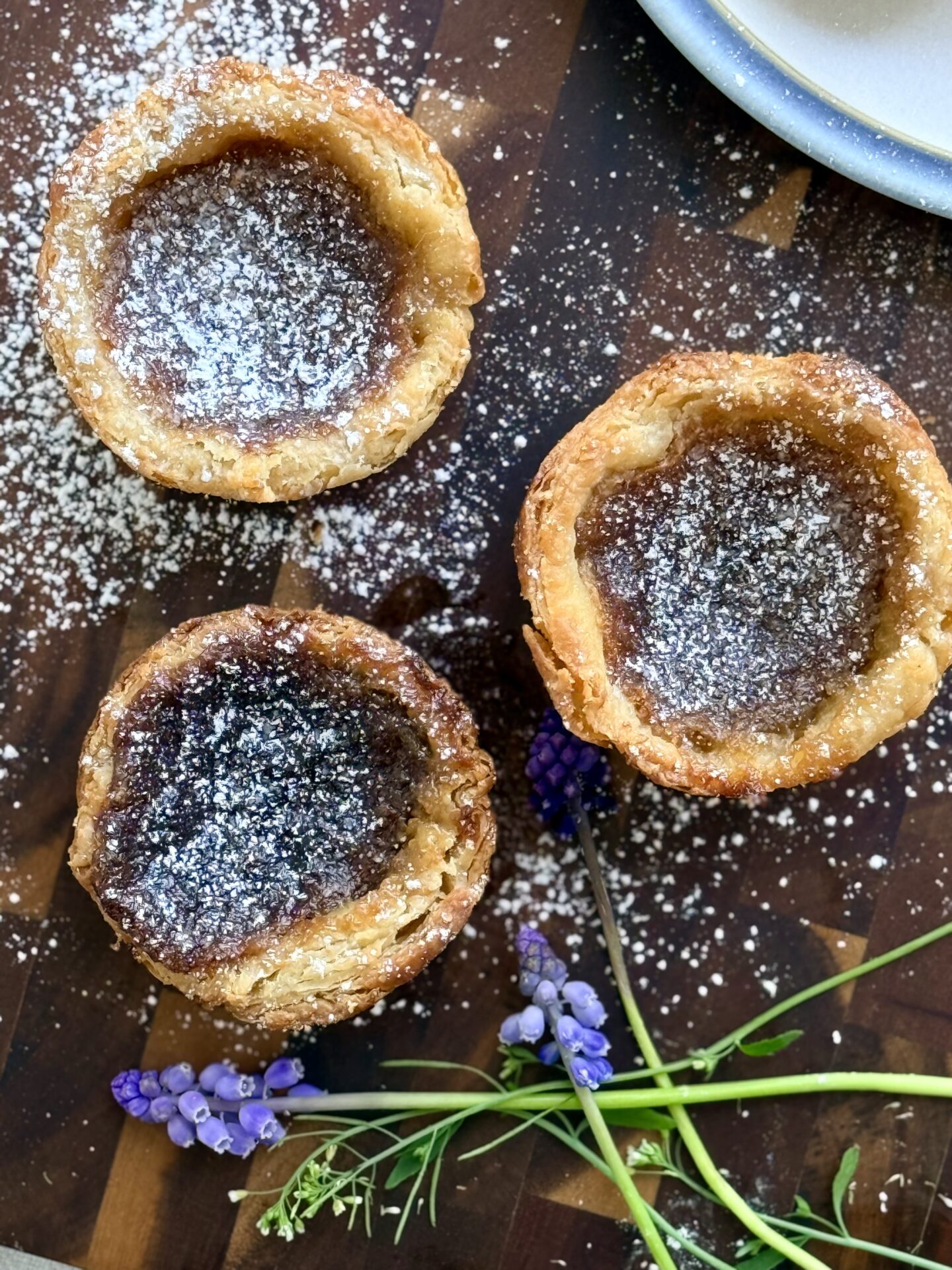 The Lakehouse Butter Tarts are seen from above, dusted in powdered sugar and resting on a wooden board. Small purple and white flowers complete the image.
