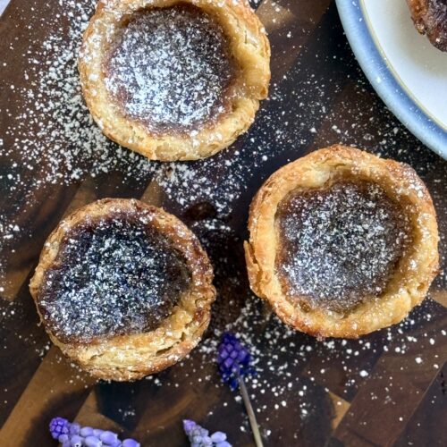 A collection of 3 freshly baked butter tarts sit on a wood board, dusted with powdered sugar and surrounded by small purple flowers.