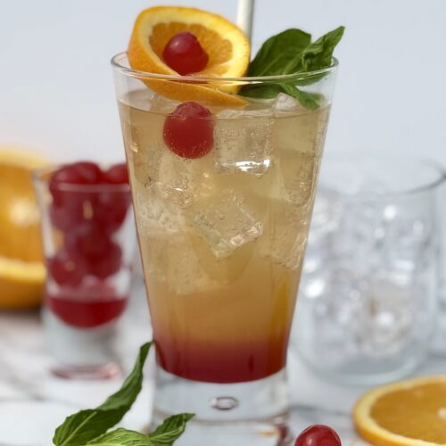 A Shirley Temple drink shows off its pretty layers of grenadine, orange juice and ginger ale, in a lavishly garnished glass.