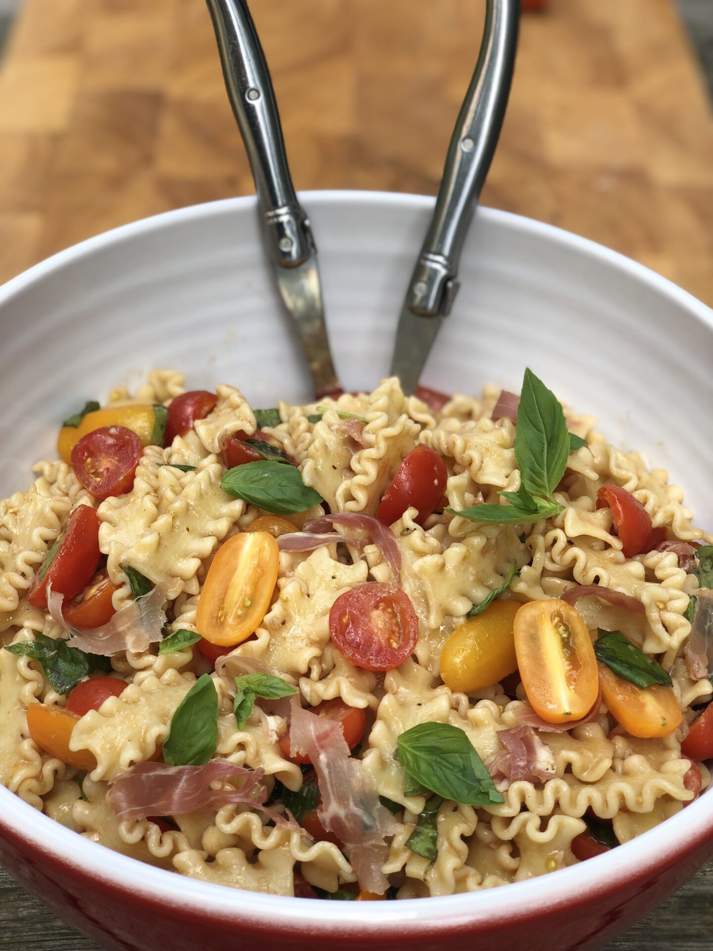 A large bowl of summer fresh pasta salad with prosciutto, cherry tomatoes and fresh basil is seen with a pair of salad servers.