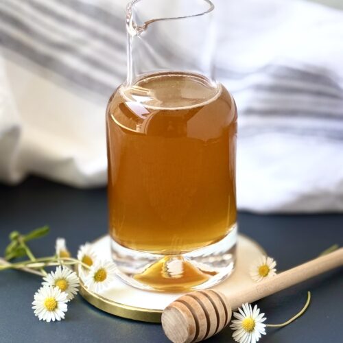 A glass bottle of honey simple syrup sits on a white and gold coaster, surrounded by tiny daisies and a honey dipper.