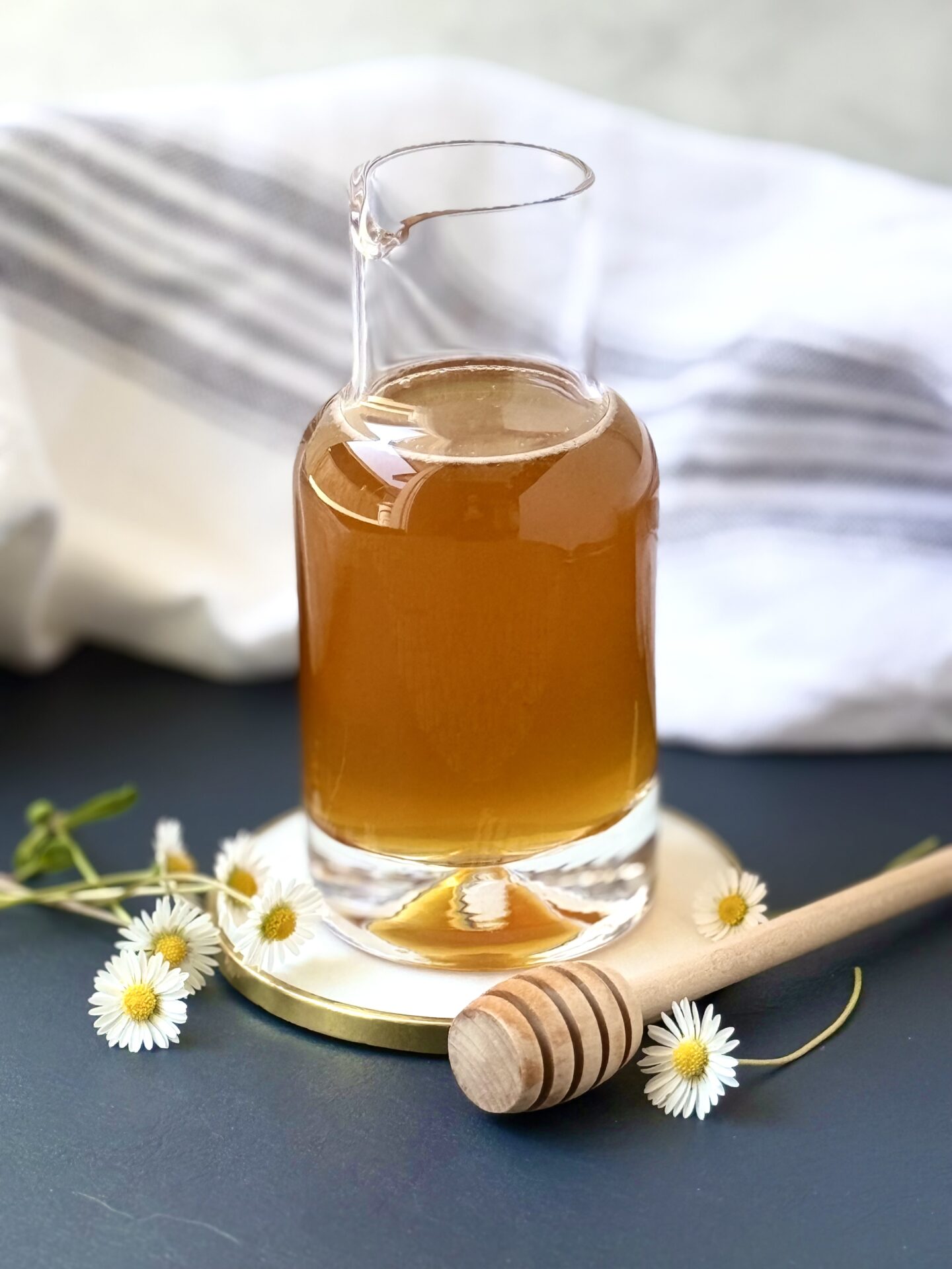 A glass bottle of amber-coloured Honey Syrup is surrounded by tiny daisies and a honey dipper.