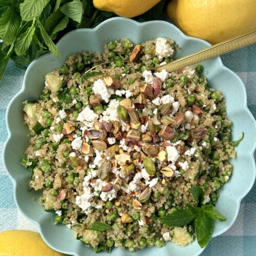 A bowl of Lemony Quinoa Salad with Spring Peas and Pistachios is seen from above, surrounded by fresh mint and lemons.