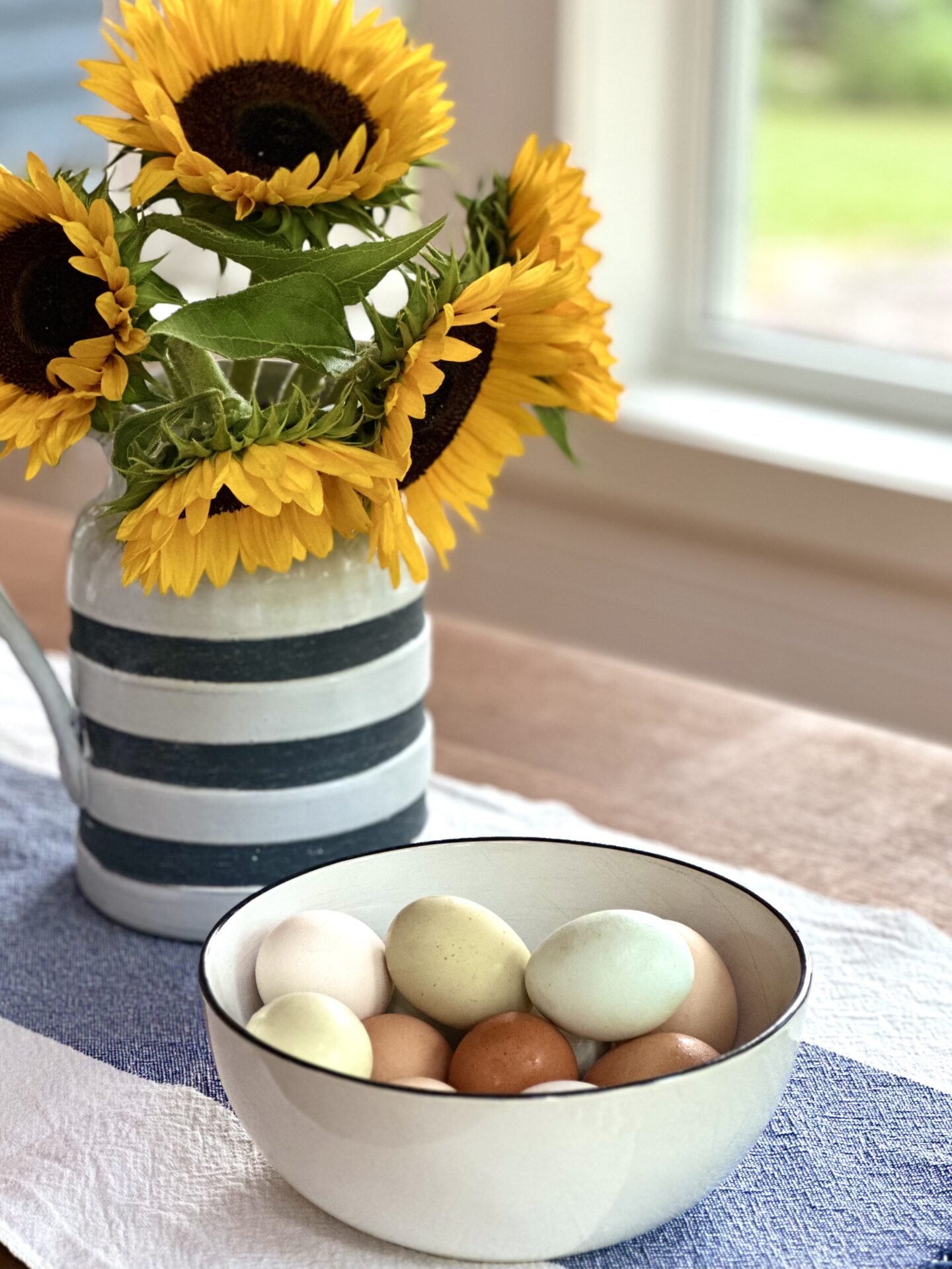 A bowl of pasted coloured eggs sits on a cottage kitchen table. A pitcher of yellow sunflowers is seen in the background.