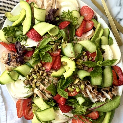 A large platter filled with Strawberry Cucumber Salad with Grilled Lemon Chicken is seen from above. The salad is beautifully arranged to show off its cucumber curls, bright red berries, creamy chunks of fresh mozzarella cheese and sprinkling of crunchy pistachios. A pair of gold-coloured salad servers sits nearby.