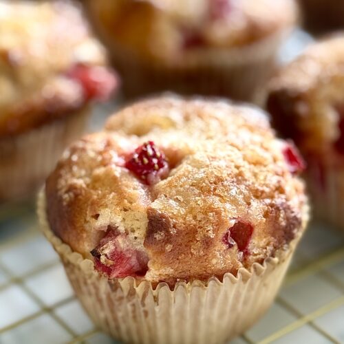 A freshly baked strawberry yogurt muffin is seen close up, showing its crispy lemon sugar coated top, and pieces of juicy strawberry.