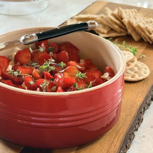 A red bowl filled with Whipped Brie with Strawberries and Fresh Thyme sits on a wood board surrounded by crackers.