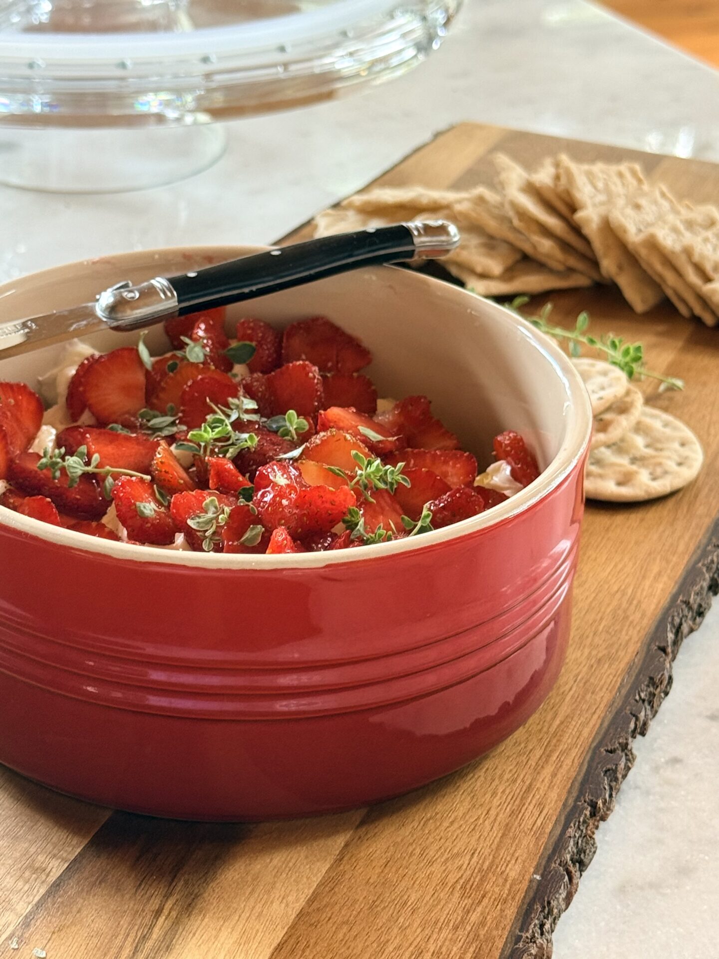 A red bowl filled with Whipped Brie with Strawberries and Thyme sits on a wood board with crackers