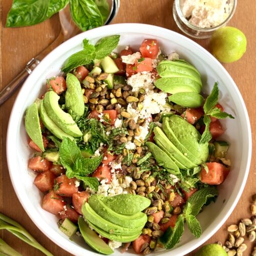 A large bowl of watermelon feta salad with avocado, pistachios and mint is seen from above, surrounded by fresh vegetables, fruit and herbs.