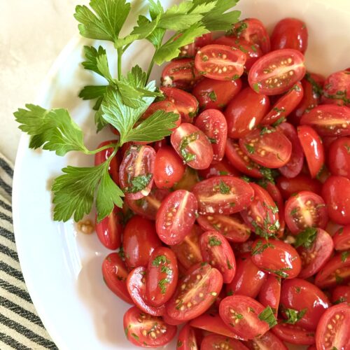 A bowl of marinated cherry tomatoes sits on a white marble countertop, surrounded by fresh herbs.