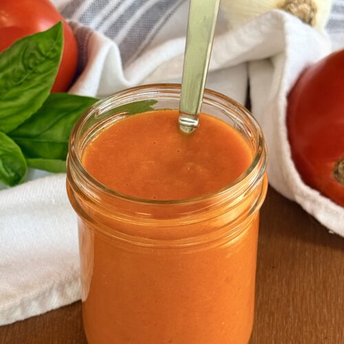 A jar of bright orange fresh tomato sauce is seen on a table, surrounded by fresh tomatoes, garlic and basil.