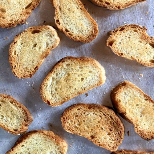 Crostini are seen from above on a baking sheet, fresh from the oven.