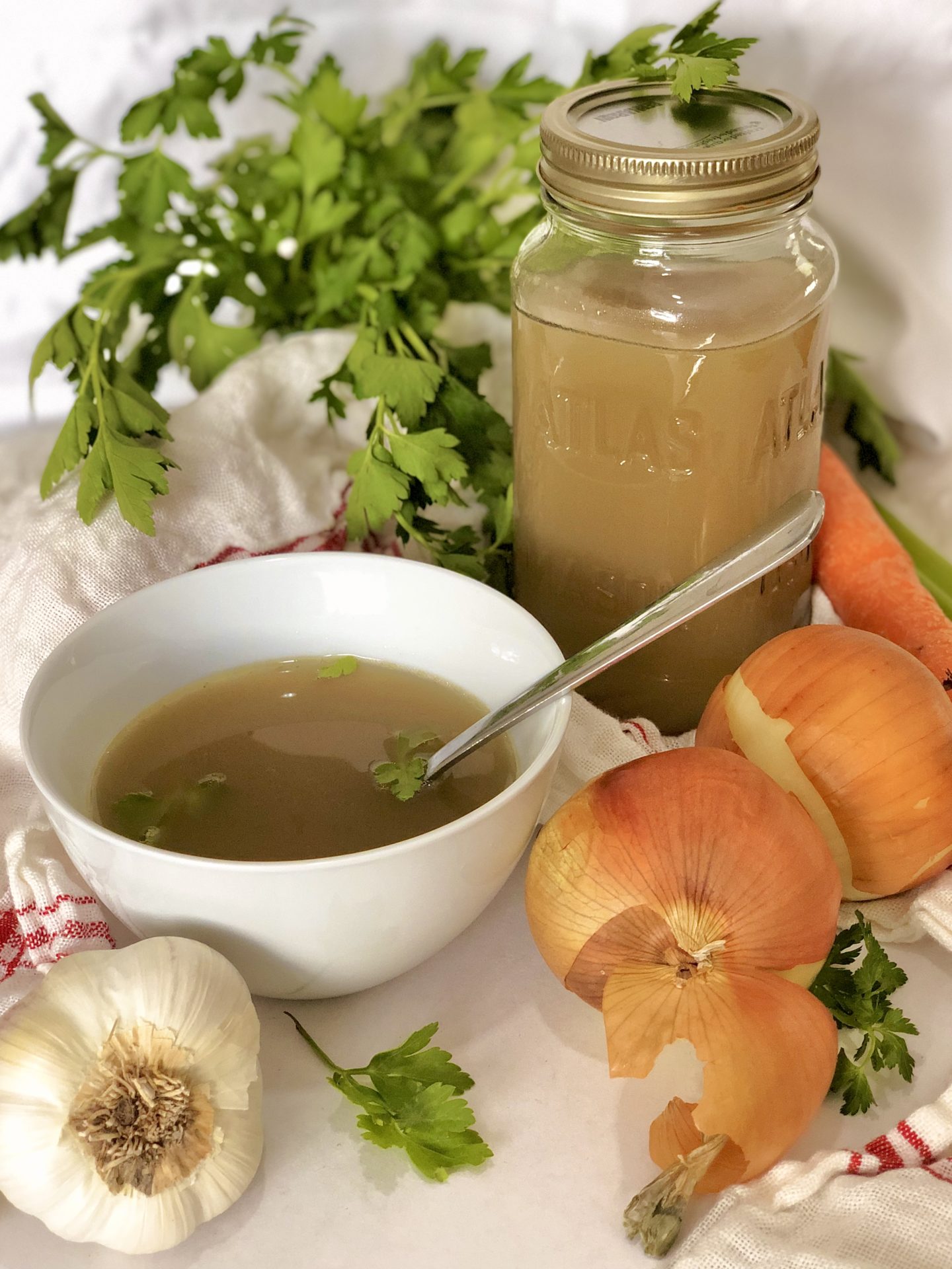 A bowl of homemade golden chicken stock sits on a white table, surrounded by fresh vegetables and herbs