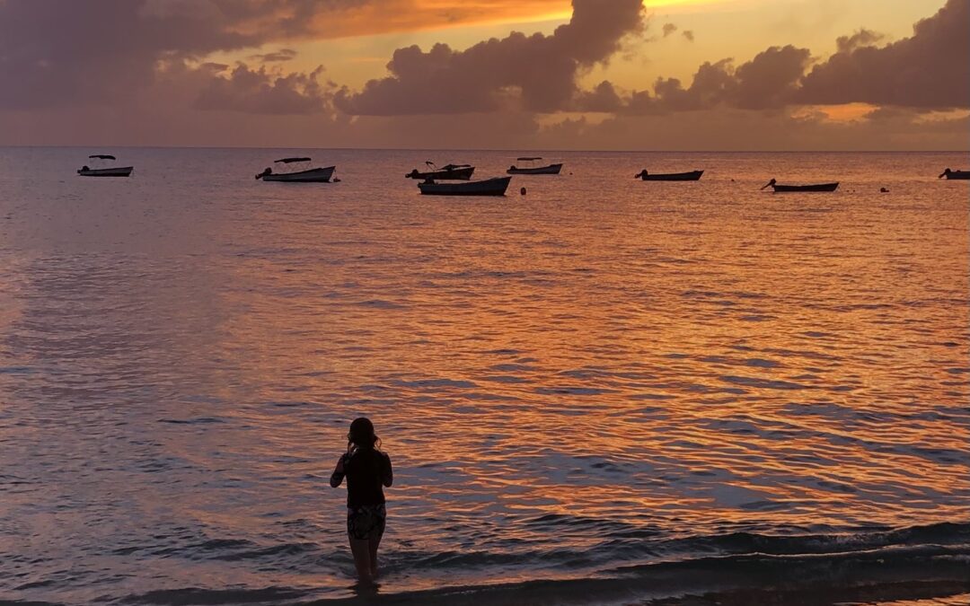 Stunning sunset on a Barbados beach with boats bobbing in the background and woman emerging from a swim in the ocean