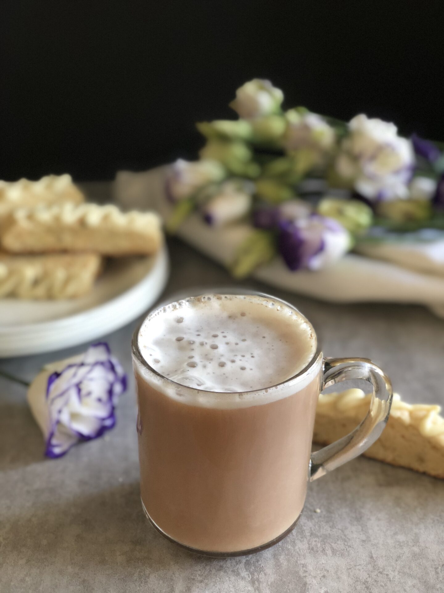 A clean mug of London Fog tea sits on a table surrounded by a platter of biscotti and some white and lavender roses 