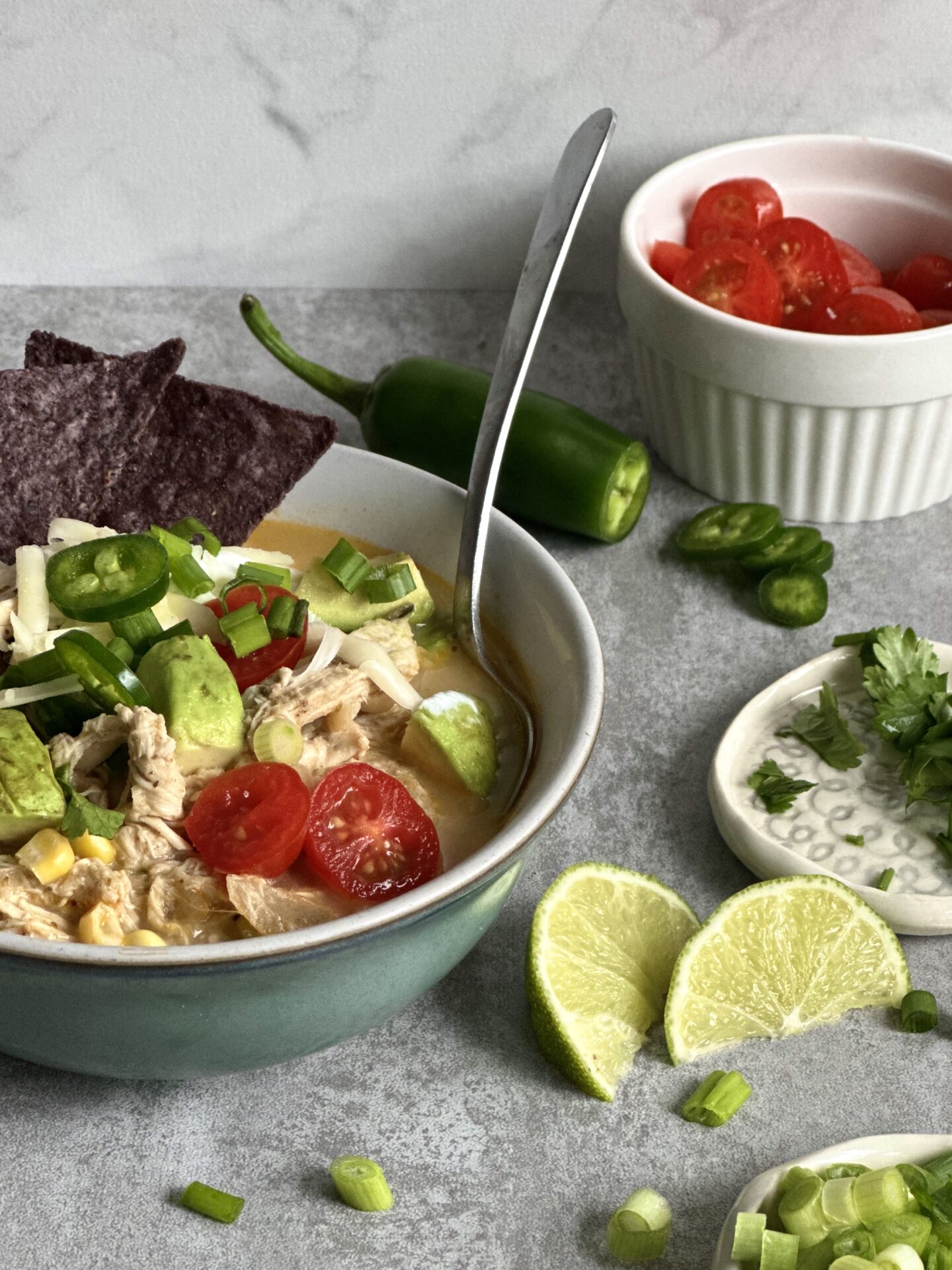 Heaping bowlful of White Chicken Chili surrounded by chopped tomatoes, scallions, blue corn chips, fresh jalapeños and sliced limes