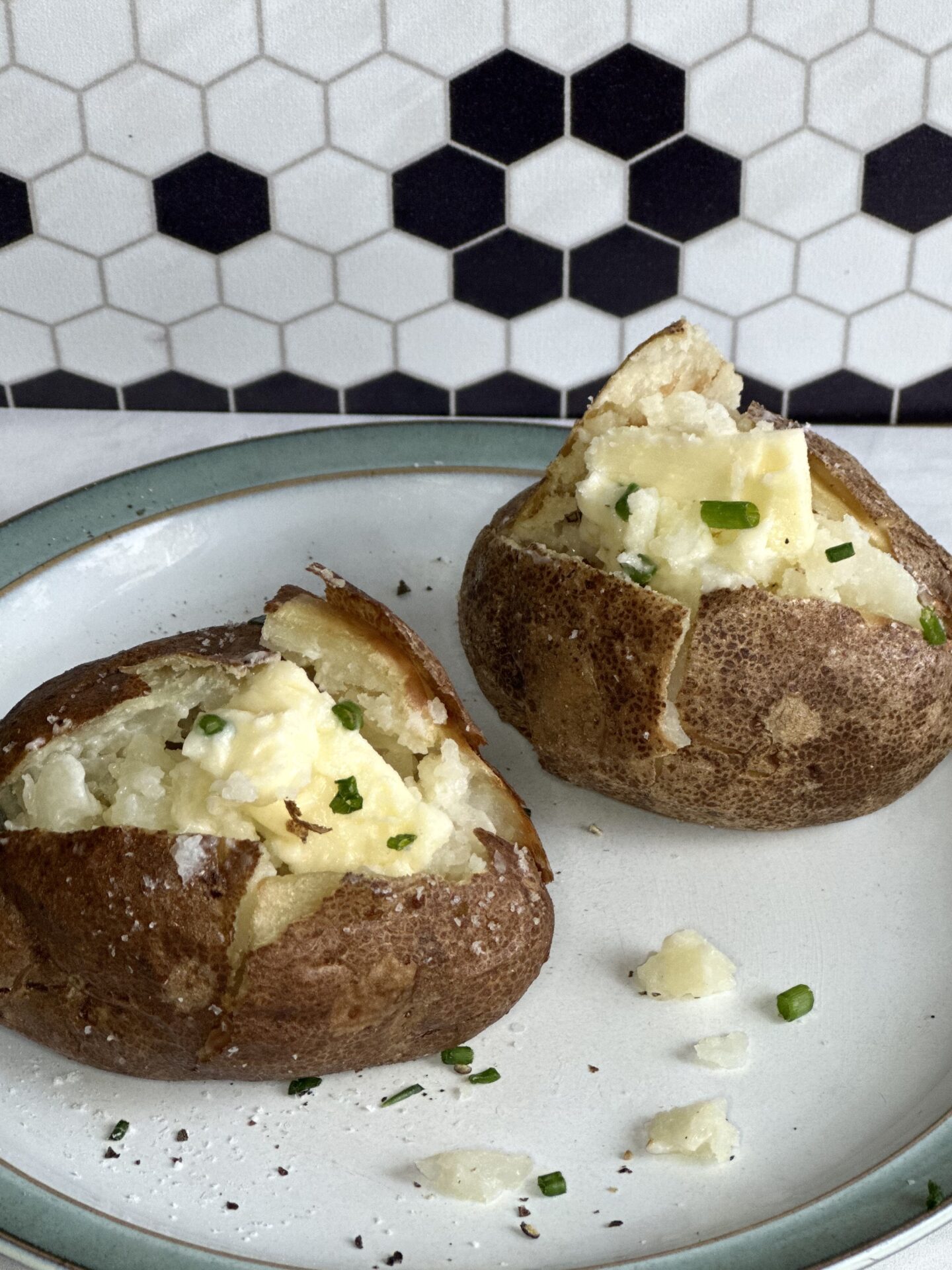 Plate of 2 perfect baked potatoes, split open to reveal a fluffy interior.  Potatoes are topped with pats of butter and sprinkles of chives.  Plate is sitting on a white marble counter top with a black and white mosaic backsplash seen in the background.