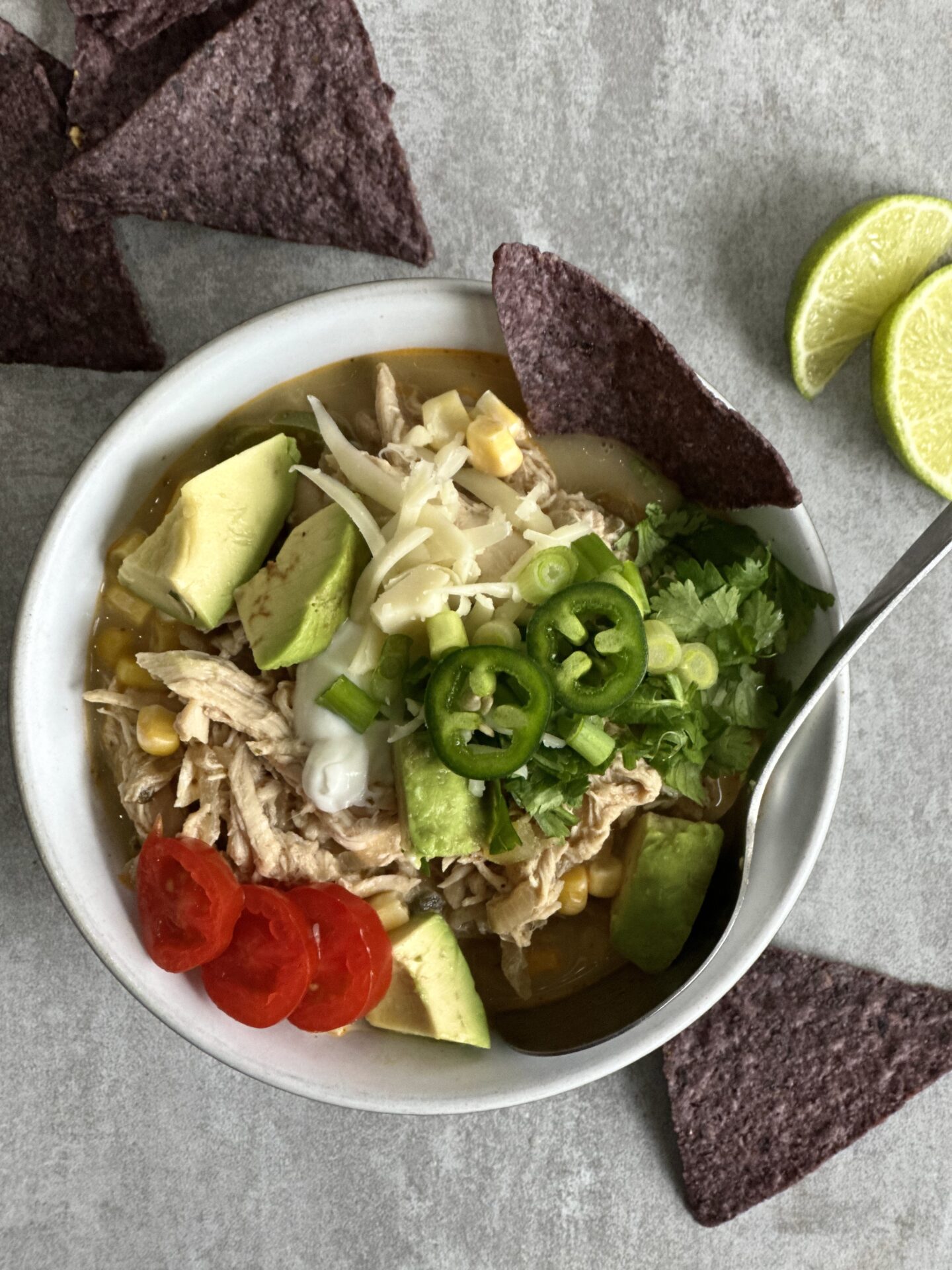 Bowl of White Chicken Chili seen from above, loaded with toppings of shredded cheese, diced avocado, chopped tomatoes, cilantro, jalapenos and green onions.  Served with lime wedges for spritzing and blue corn tortilla chips.