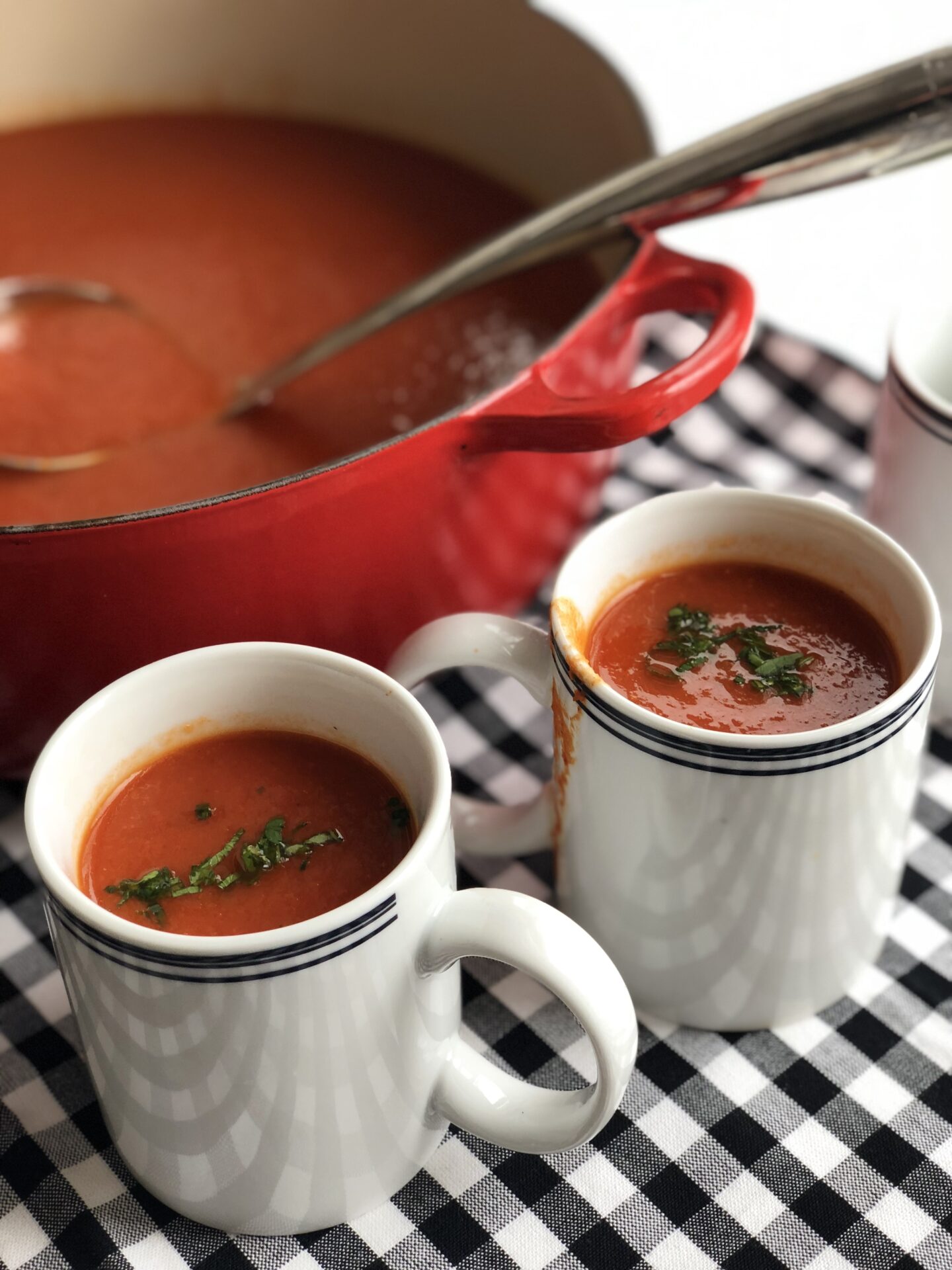 A red enamelled pot of tomato soup with a ladle and two mugs, on a black and white gingham tablecloth