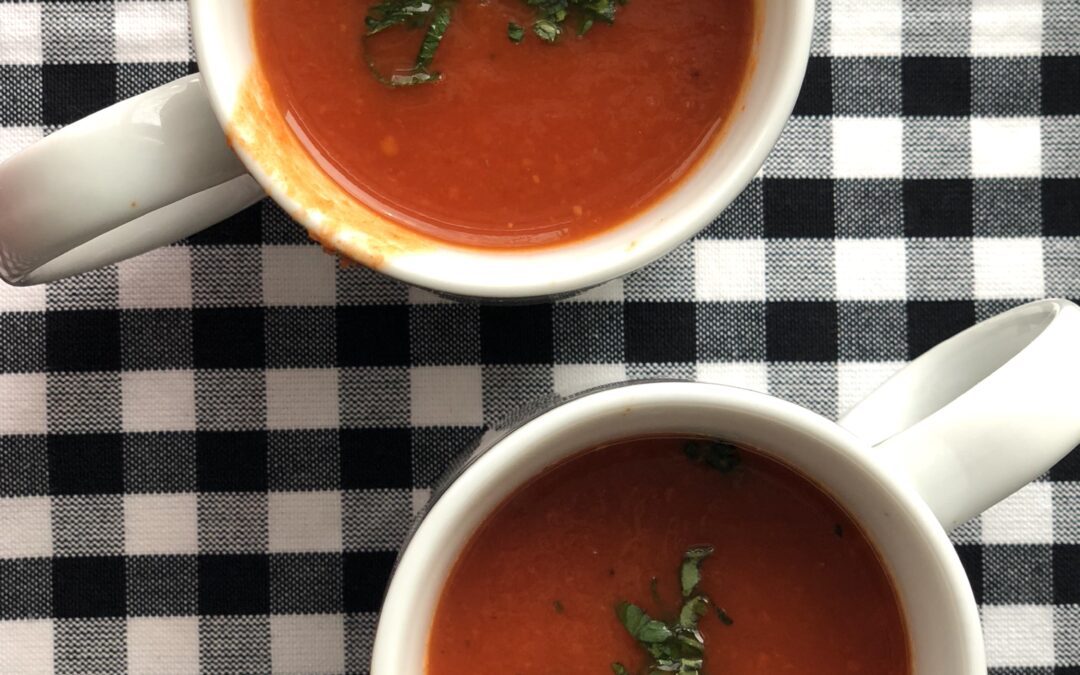 Two mugs of homemade tomato soup, set on a black and white checkered table cloth, seen from above.
