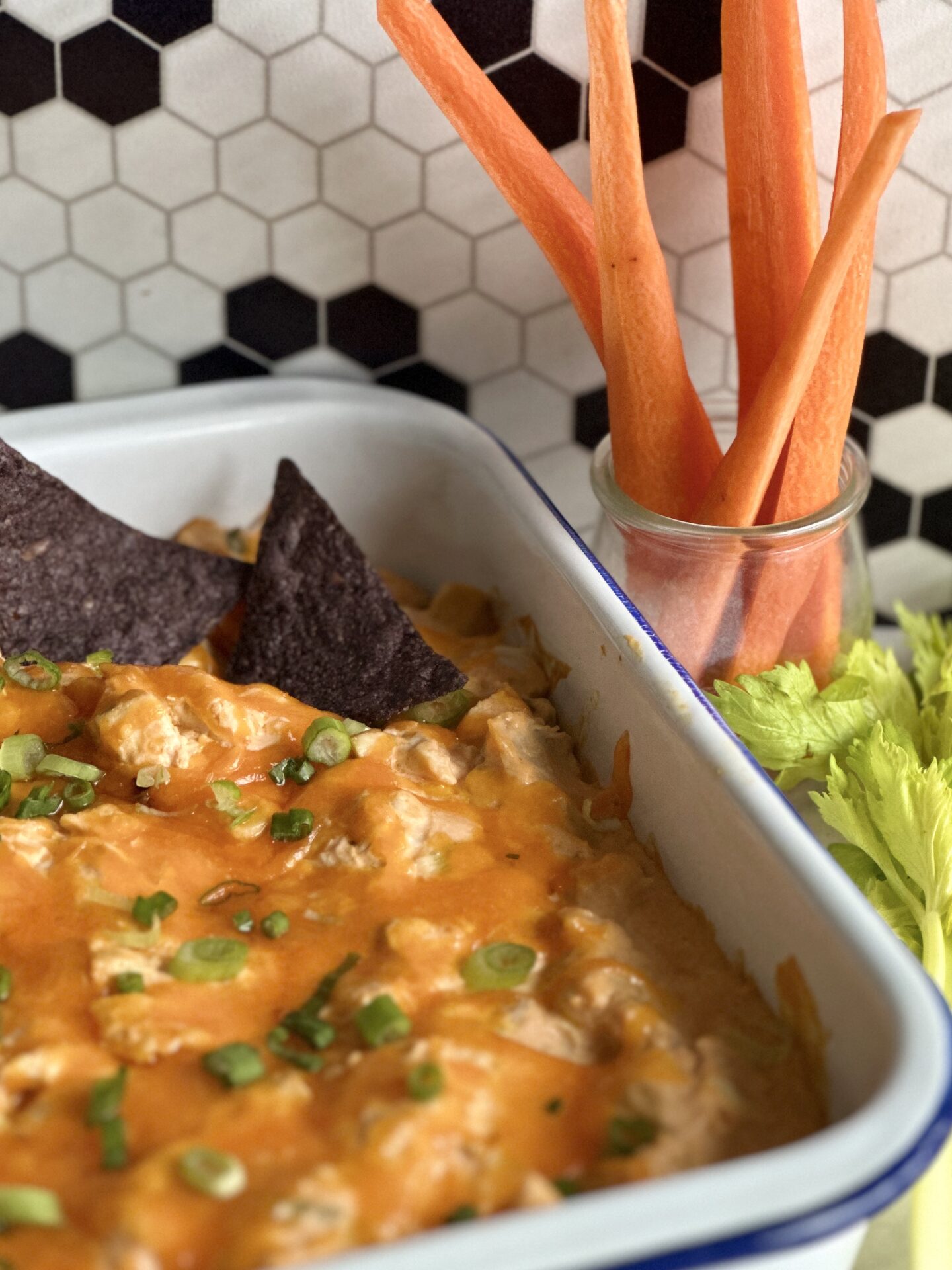 White enamel pan of Hot and Spicy Buffalo Chicken Dip with a topping of melted cheddar and green onions.  Seen on a counter top with vegetables and blue corn chips for dipping.
