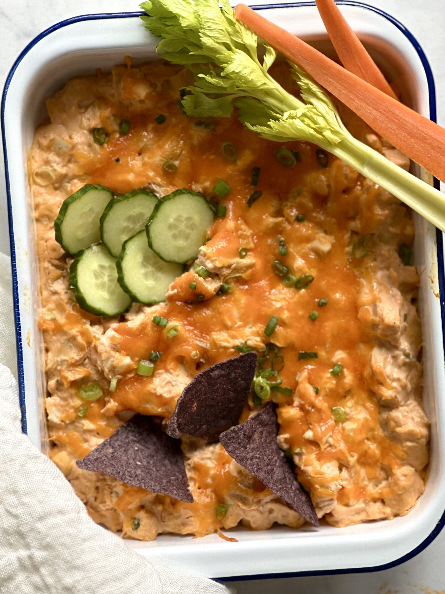 White enamel pan of Hot and Spicy Buffalo Chicken Dip with blue corn chips and vegetables for scooping.