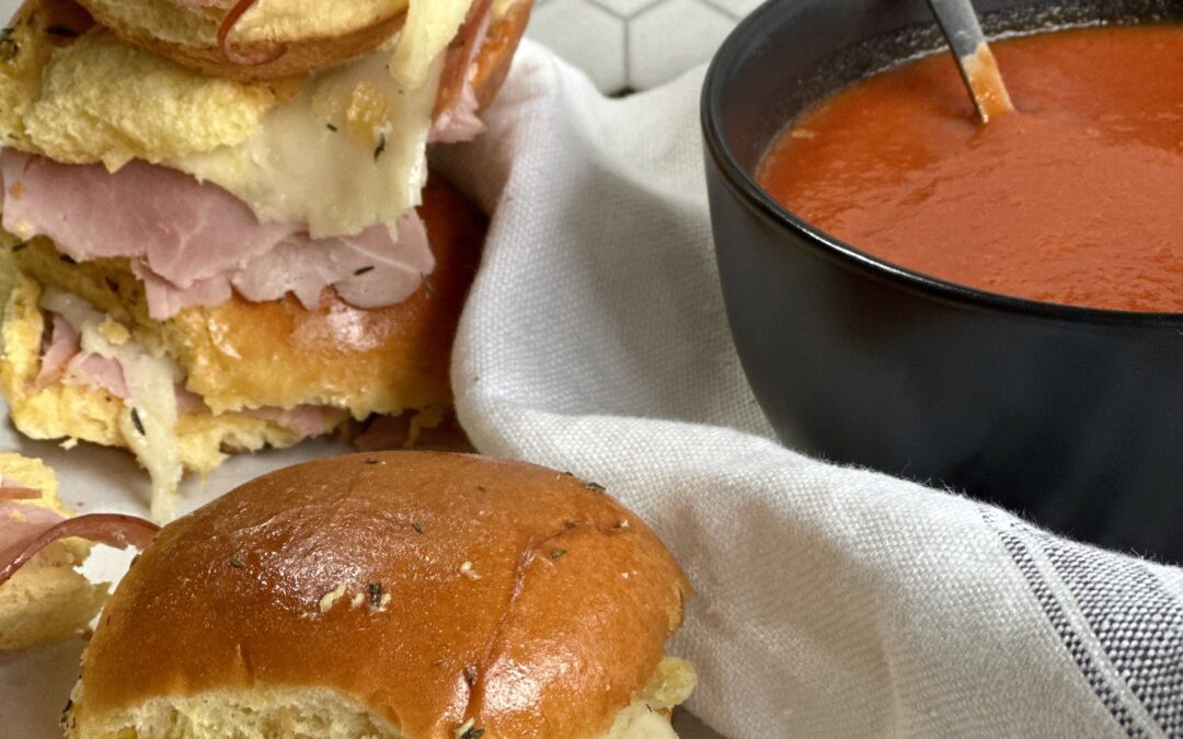 Hot and melty ham and cheese sliders stacked up beside a bowl of homemade tomato soup