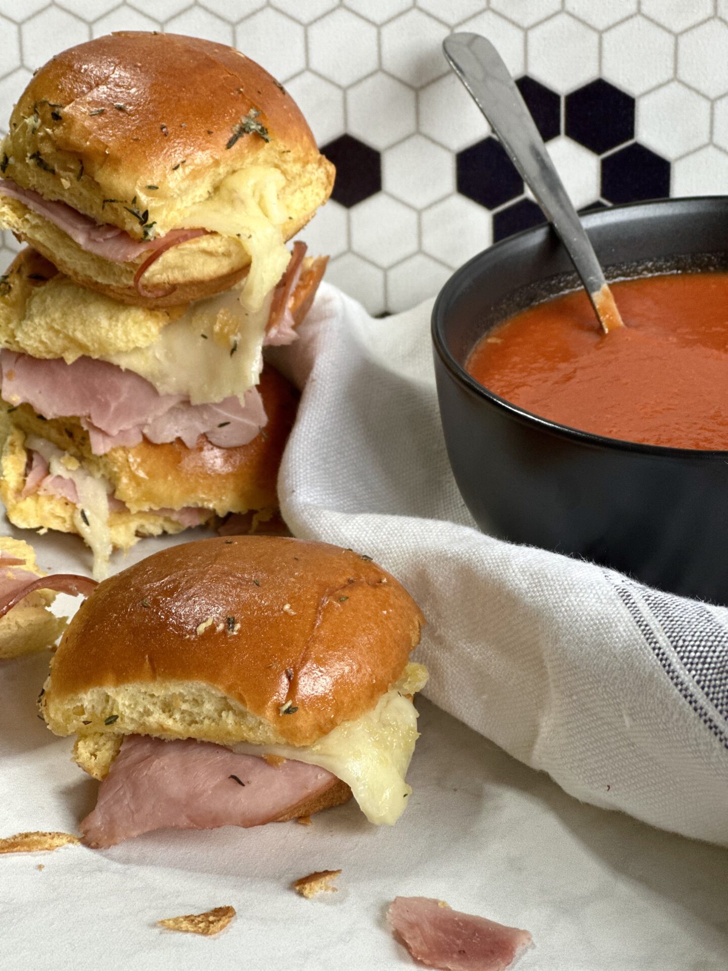 Hot and melty ham and cheese sliders stacked up beside a bowl of homemade tomato soup