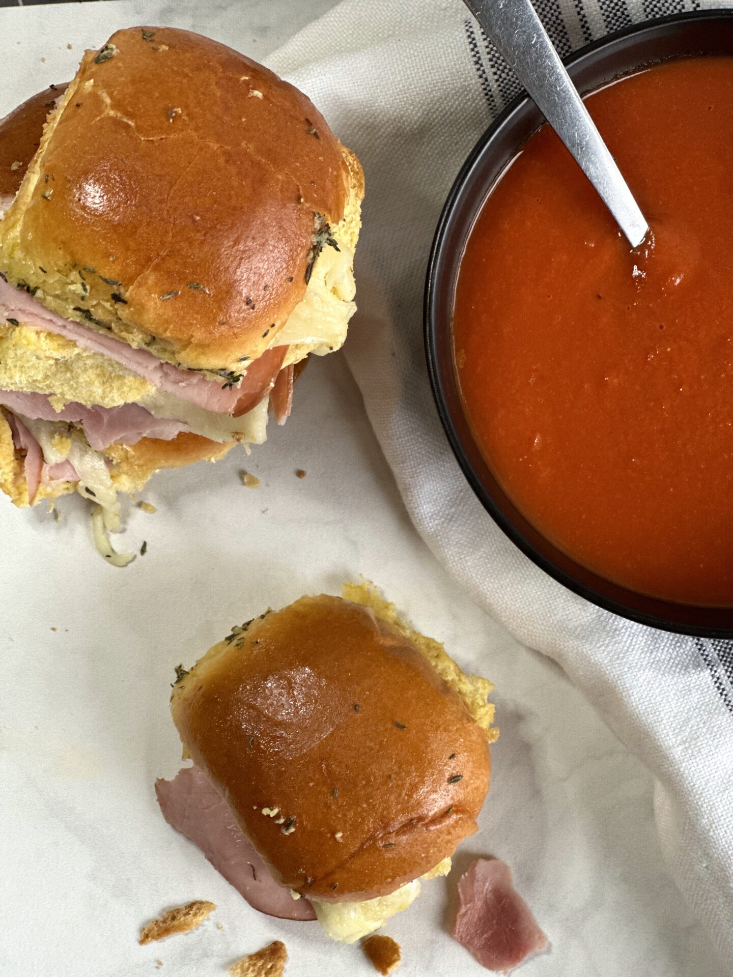 Hot ham and cheese sliders on golden buns seen from above with a bowl of homemade tomato soup.