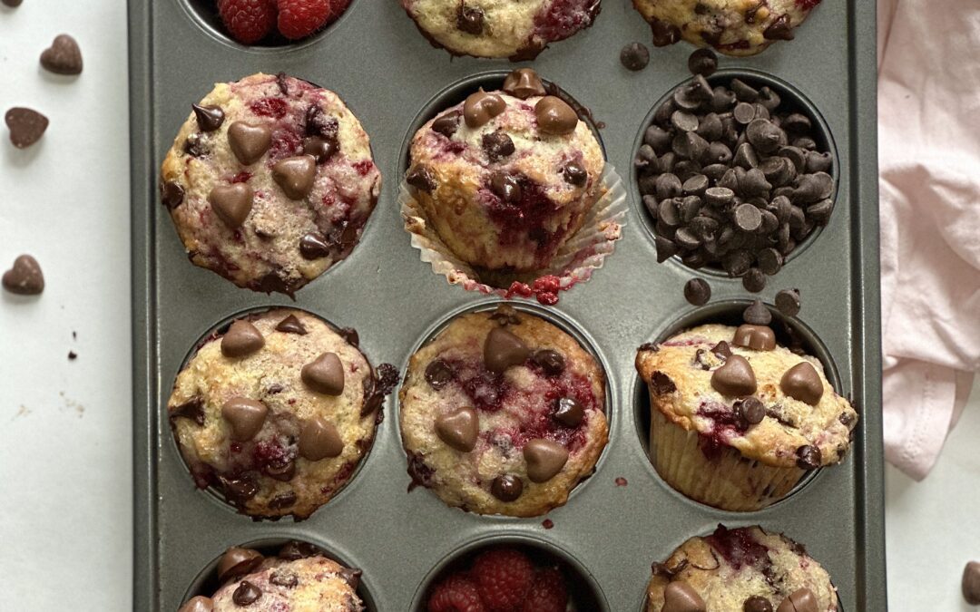 Muffin pan full of decadent raspberry chocolate muffins surrounded by fresh raspberries and heart shaped chocolates