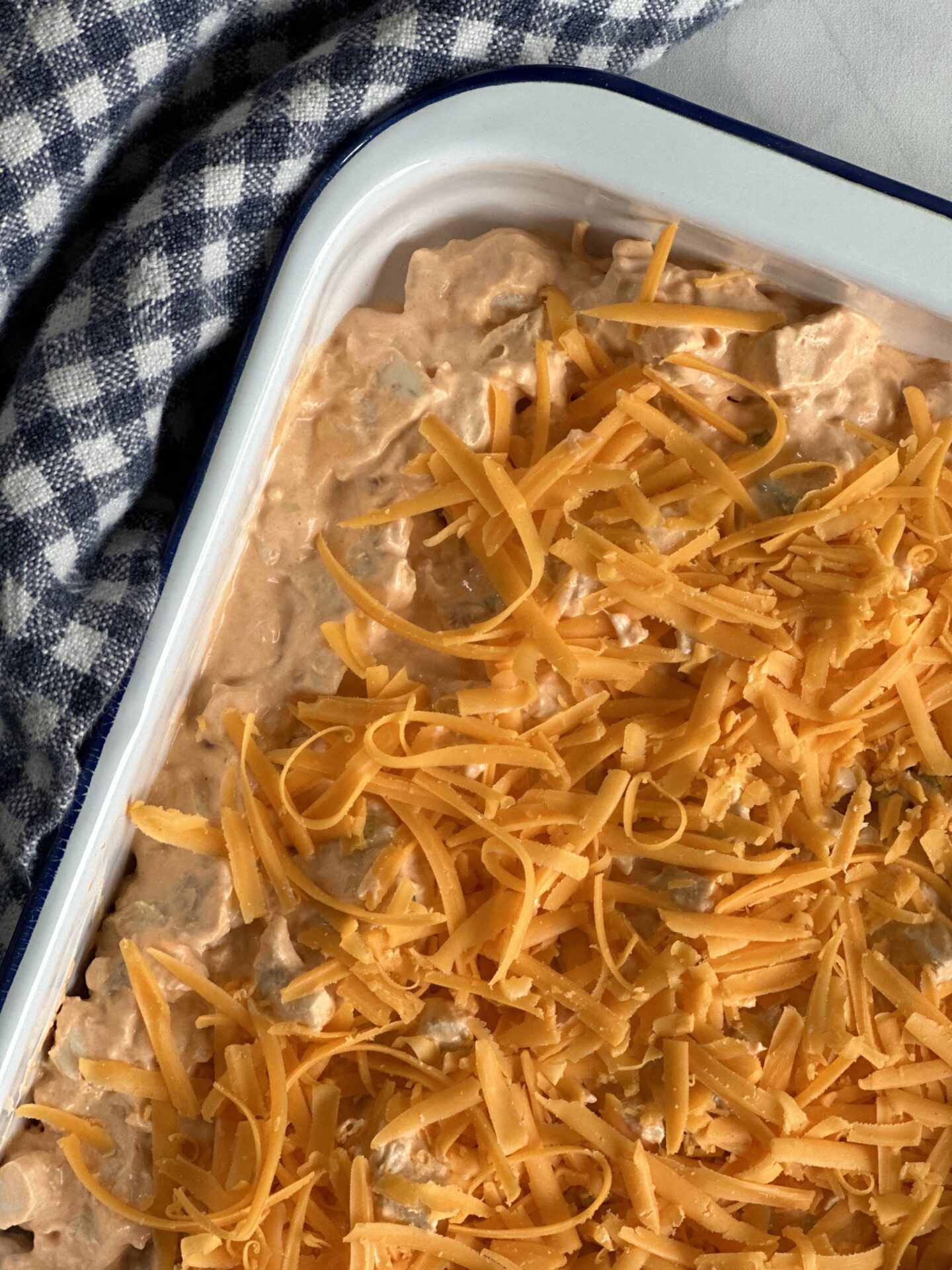 White enamel pan with Hot and Spicy Buffalo Chicken Dip, topped with grated cheddar cheese, ready to be warmed in the oven.