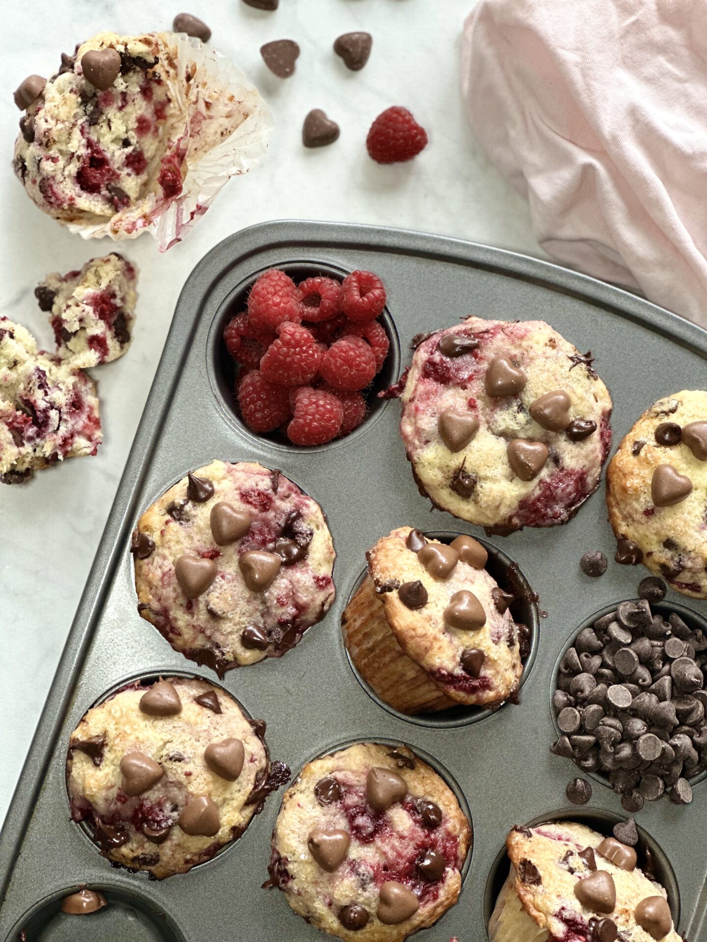 A pan of decadent raspberry chocolate chip muffins garnished with chocolate hearts for valentines day, seen with fresh raspberries and heart shaped chocolates