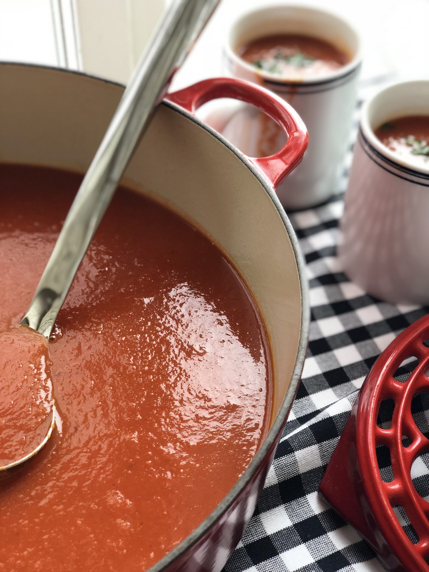 A large pot of homemade tomato soup seen from above, with a ladle and mugs for serving
