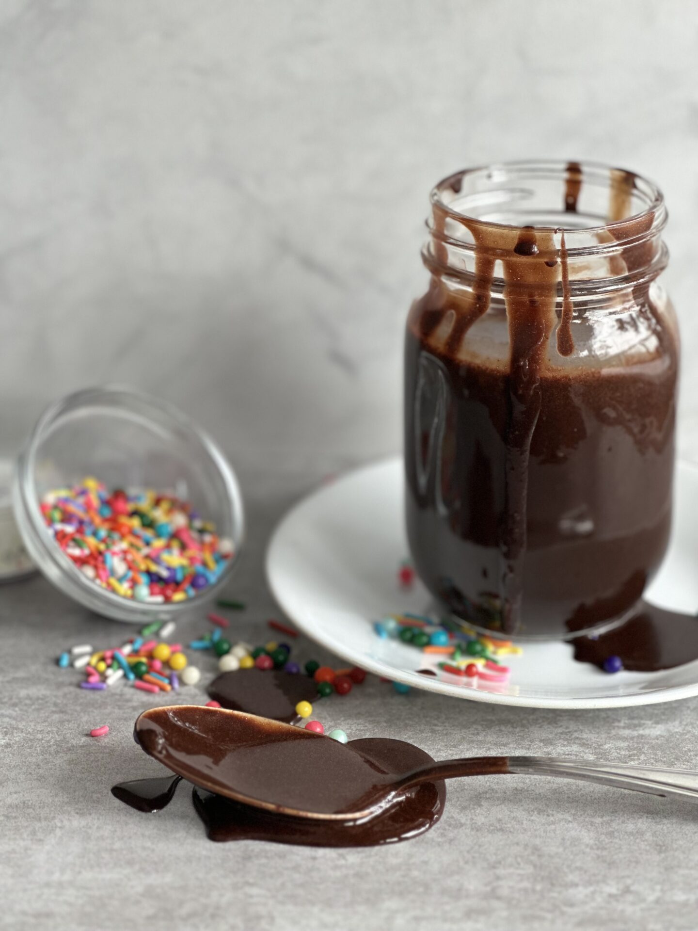 A glass jar of hot chocolate fudge sauce surrounded by drips of chocolate fudge sauce and colourful sprinkles
