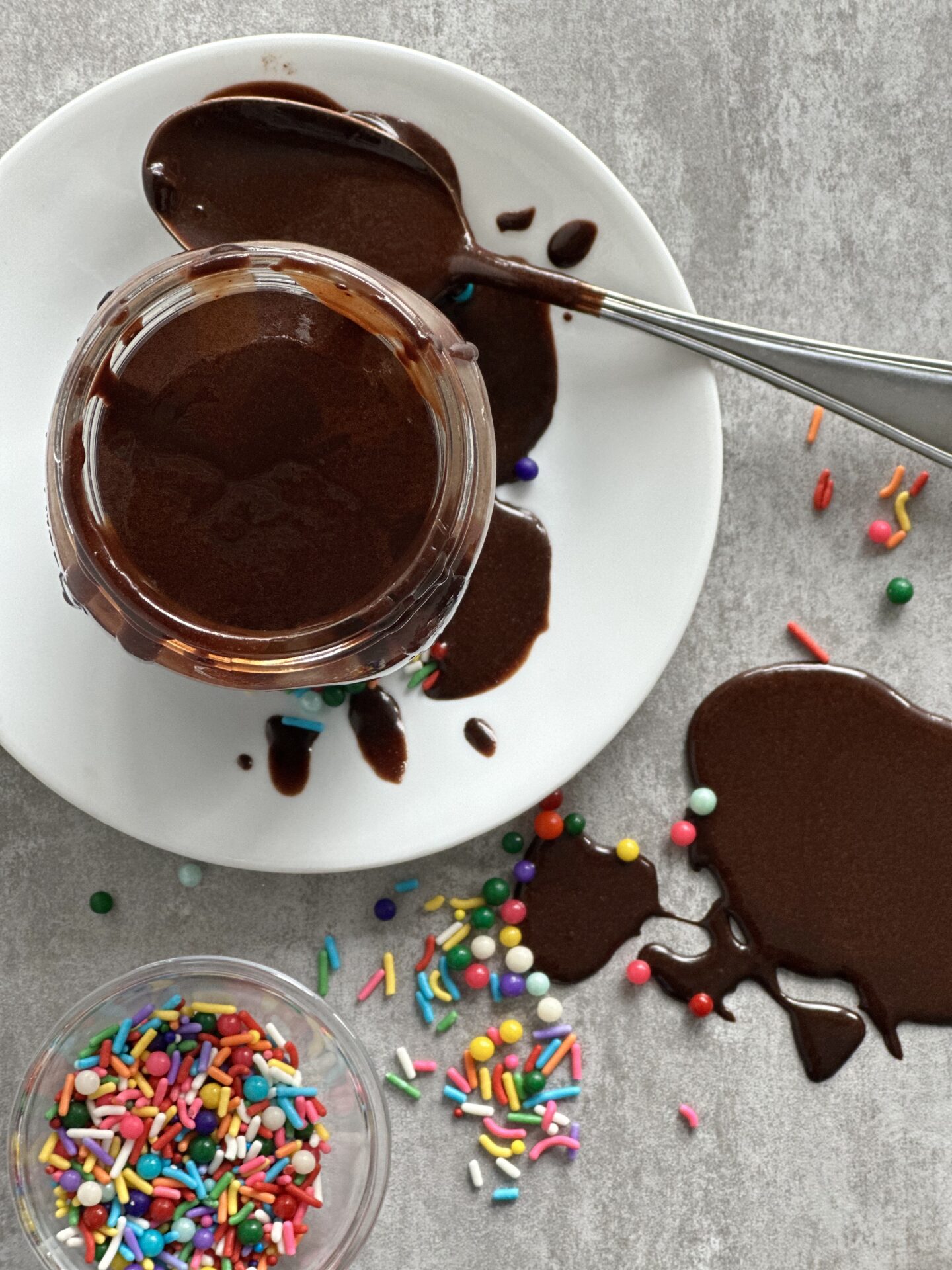 Jar of Hot Chocolate Fudge Sauce seen from above, surrounded by spilled sauce and colourful sprinkles