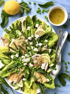 Spring salad with asparagus peas feta and roast chicken displayed on a platter surrounded by lemon mint and a bowl of dressing