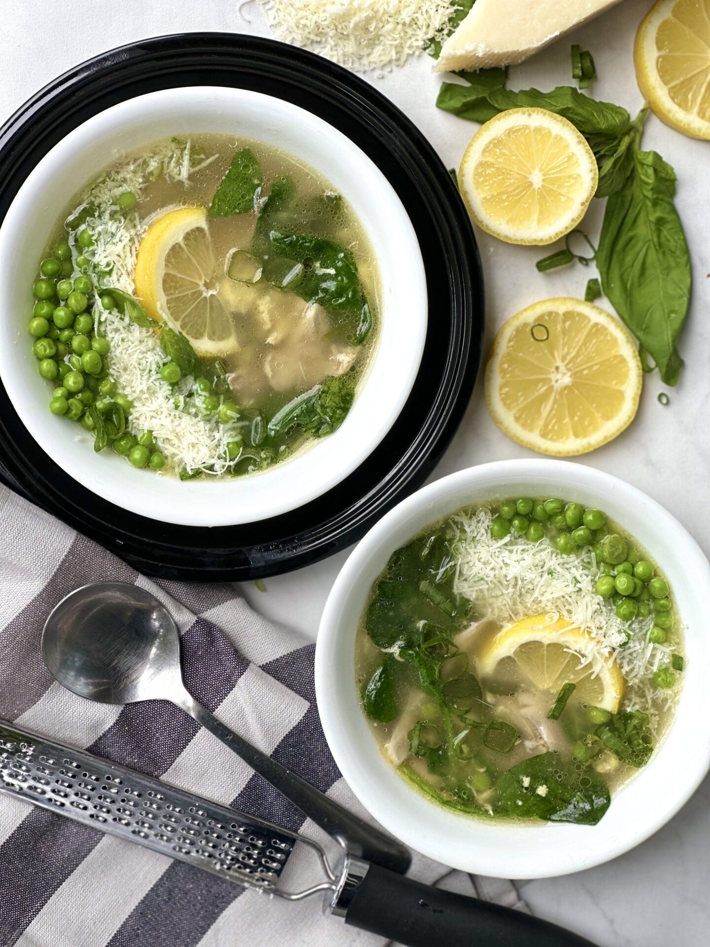 Two bowls of spring straciatella soup, seen from above garnished with fresh basil lemon peas and freshly grated parmesan cheese