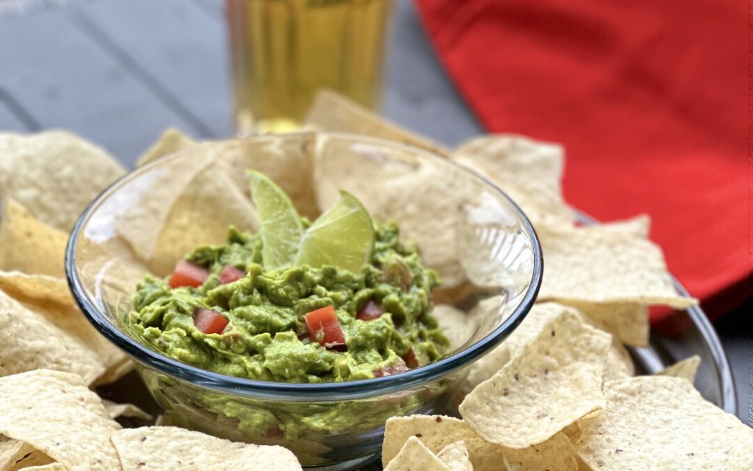 A platter of chips with fresh homemade guacamole sitting on an outdoor patio table with a glass of cold beer and a bright red napkin