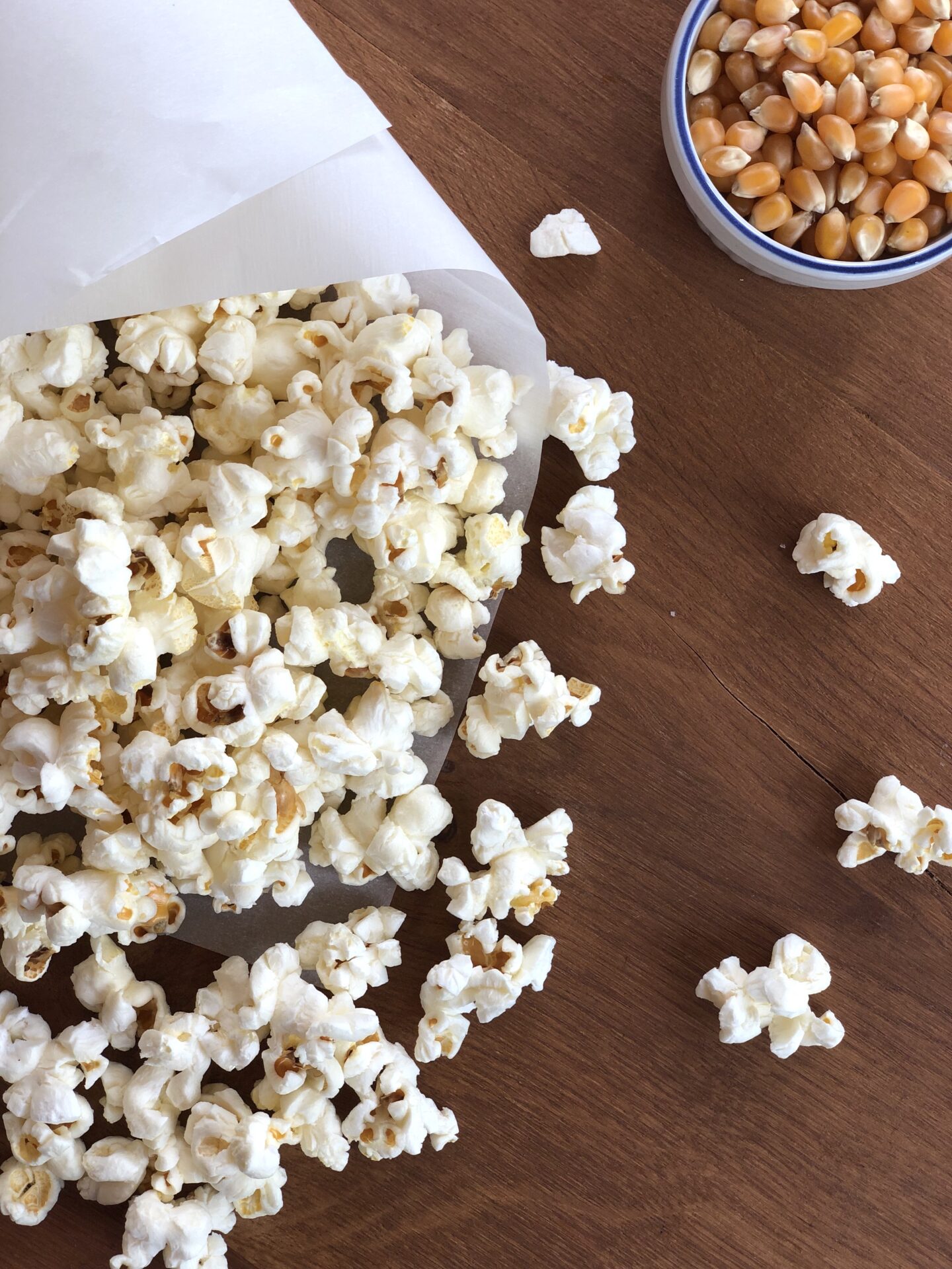 Paper cone of fresh truffle popcorn seen from above on a wood table with a dish of popcorn kernels