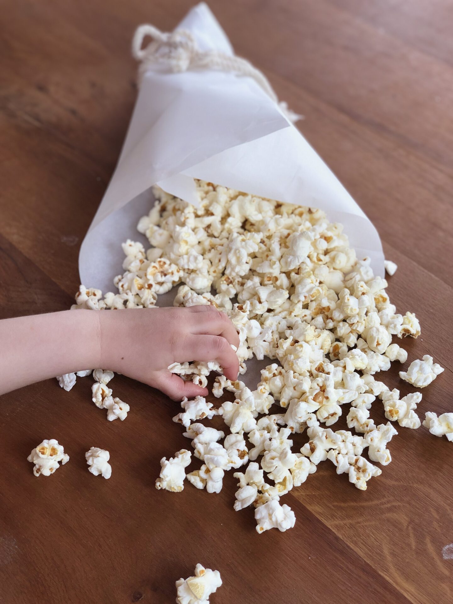 A little hand reaches into grab a handful of truffle popcorn from a paper cone of popcorn sitting on a wood table