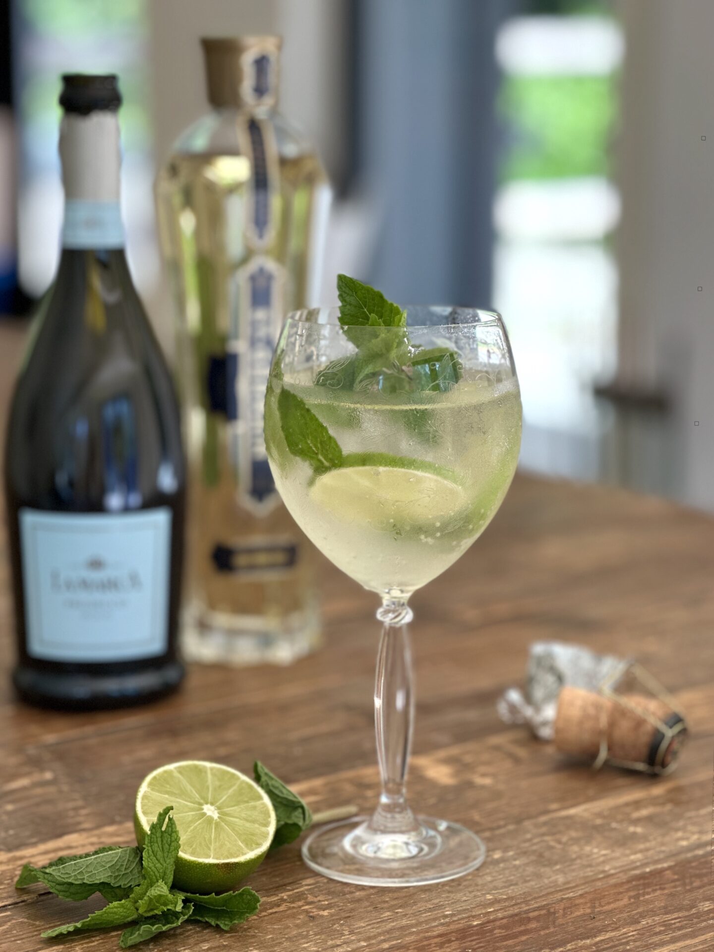 A Hugo Spritz cocktail on a wood table surrounded by its ingredients 