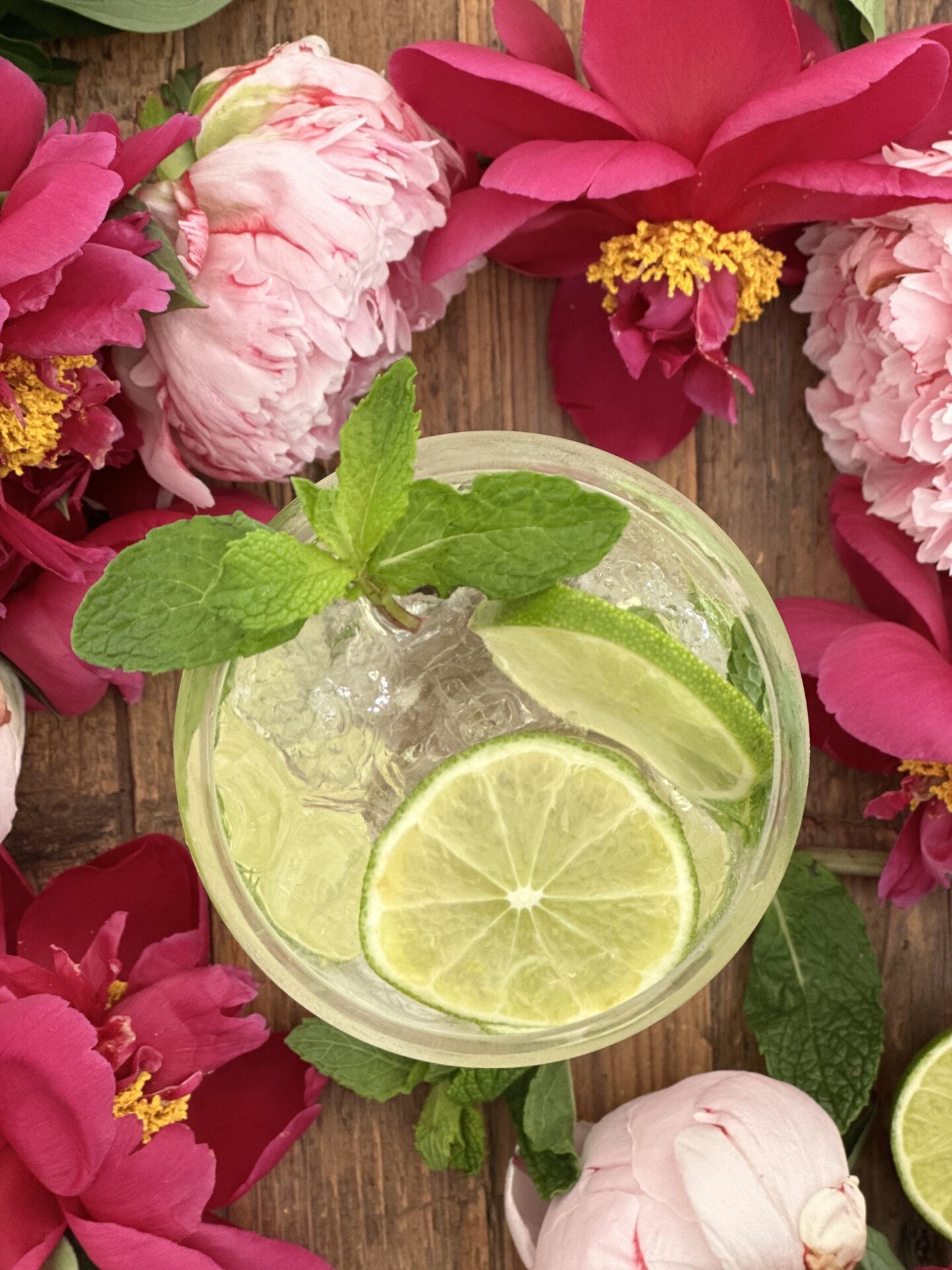Hugo Spritz cocktail seen from above with garnishes or lime wheels and fresh mint surrounded by pink and magenta peonies