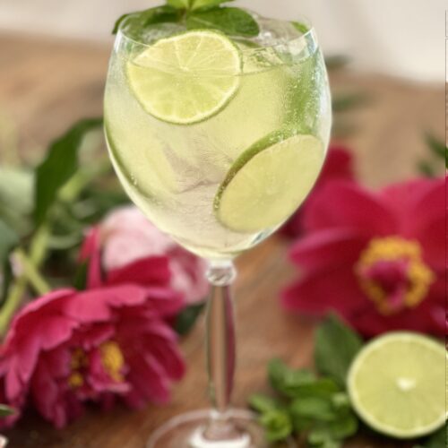 Frosted glass filled with a Hugo Spritz cocktail garnished with fresh mint and lime wheels on a wooden table surrounded by peonies