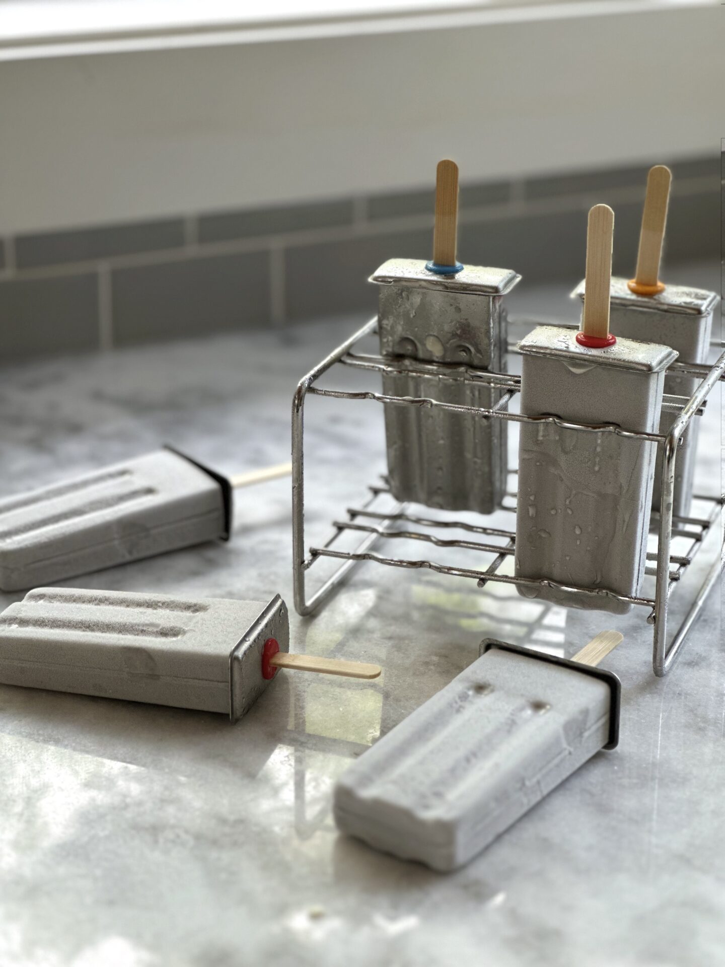 Homemade popsicles in frosty stainless steel popsicle moulds sitting on a white marble counter with grey tile backsplash in the background