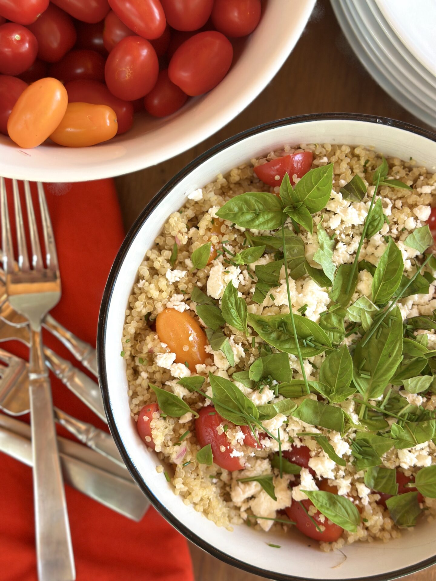 A bowl of Cherry Tomato Quinoa Salad garnished with fresh basil leaves is seen from above. A bowl of heirloom cherry tomatoes and a stack of forks are seen in the background.