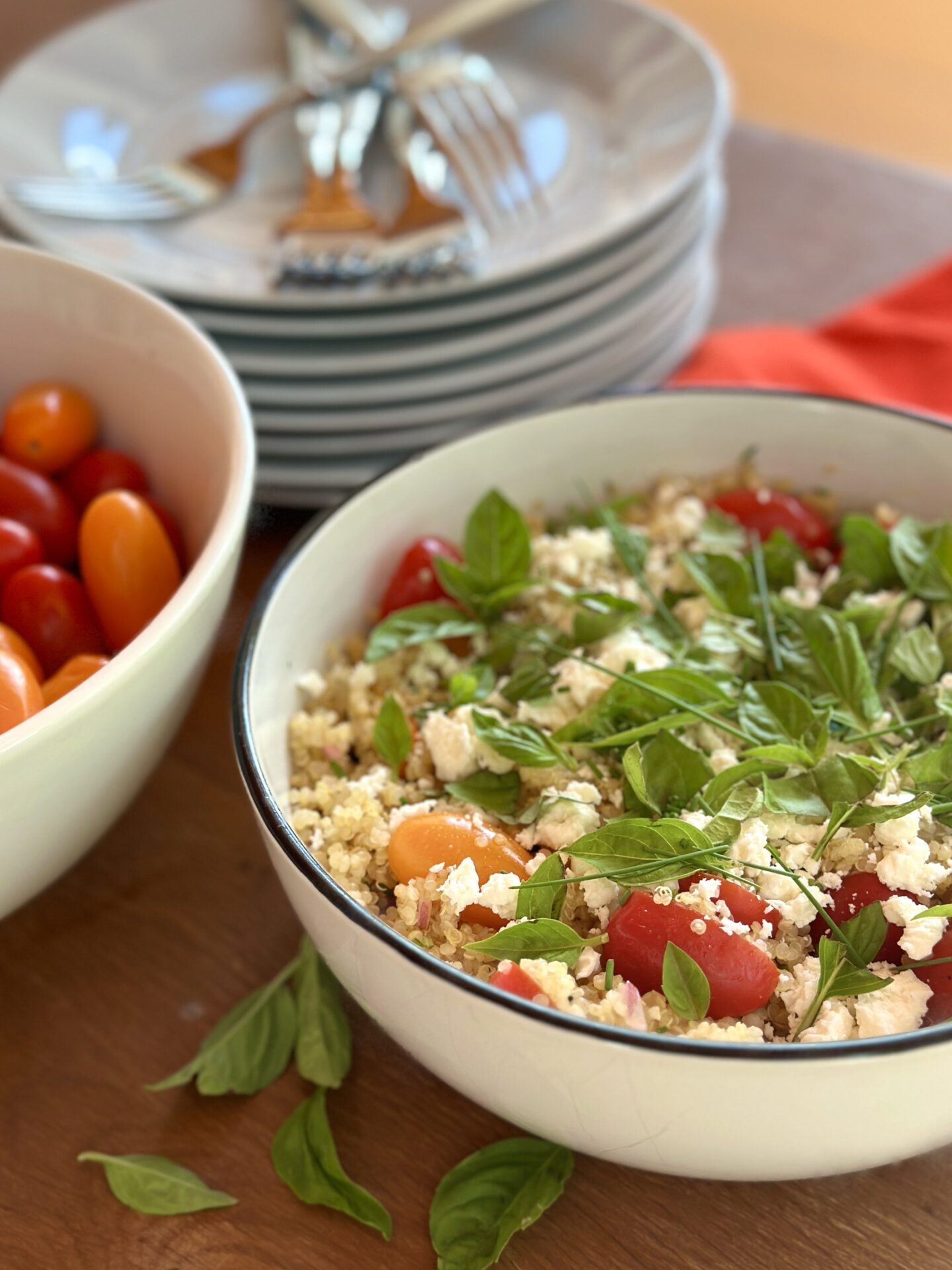 A large white bowl of cherry tomato quinoa salad garnished with fresh basil is seen, with a stack of white plates and forks in the background