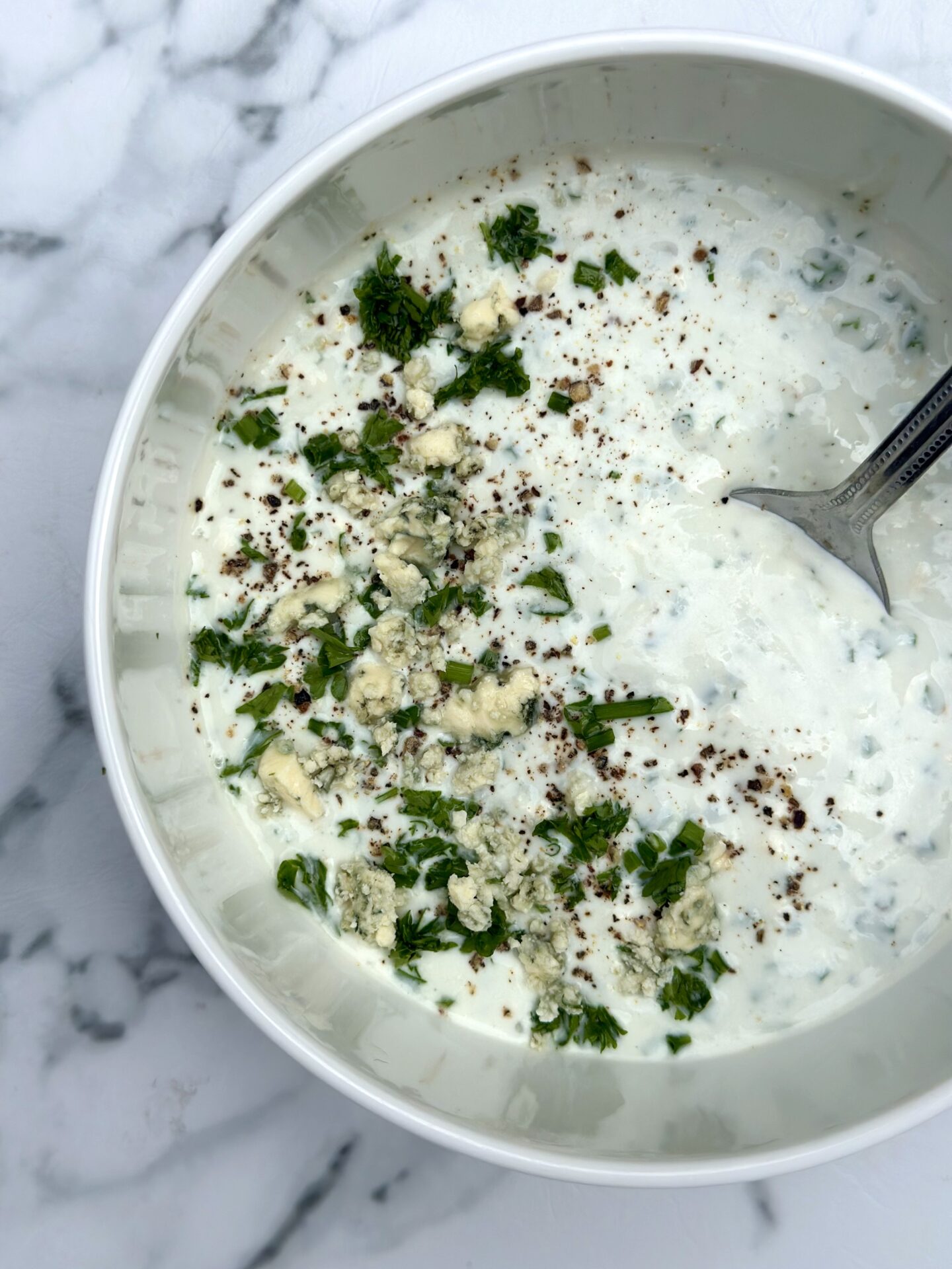 Bowl of homemade blue cheese dressing seen from above garnished with crumbled blue cheese and fresh herbs, sitting on a white marble table