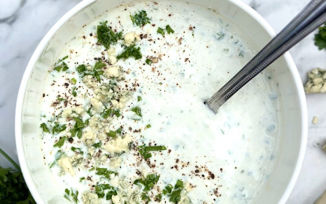 Bowl of homemade blue cheese dressing seen on a white marble countertop surrounded by lemon, garlic and fresh herbs