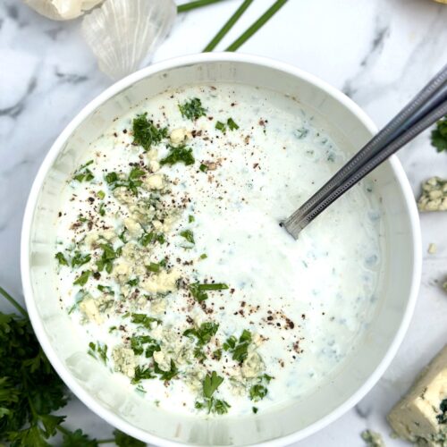 Bowl of homemade blue cheese dressing seen on a white marble countertop surrounded by lemon, garlic and fresh herbs